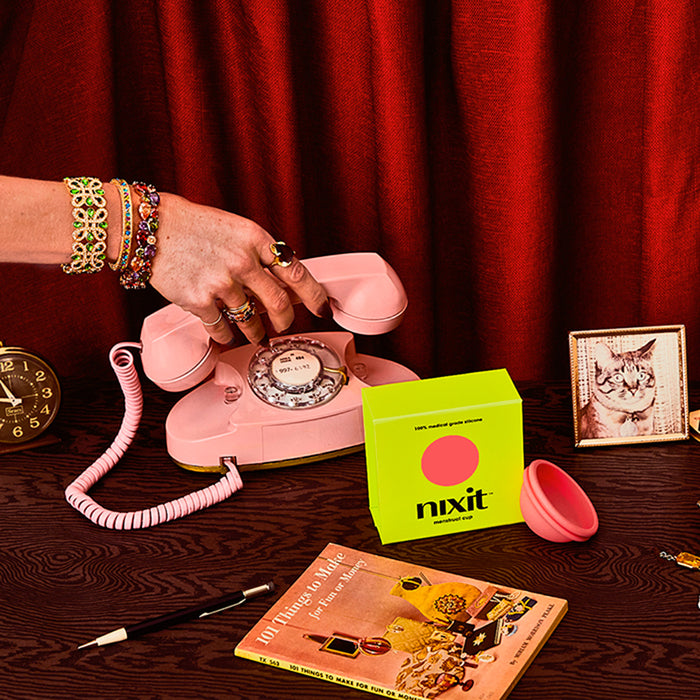 Vintage pink rotary phone with a hand wearing colorful bracelets holding the receiver, alongside a nixit menstrual cup box with a red curtain background.