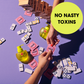 two arms extend over a messy table of dominos to exchange a condom in it's wrapper.  On the table are nixit condoms and a wooden block game, with a bottle of nixit personal lubricant.   Wording in a circle says 'no nasty toxins'